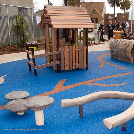 Outdoor playground with blue flooring and small wooden structure labeled "Kid's Cabin." Fake hollow log and mushrooms for stepping and crawling through scattered throughout playground.