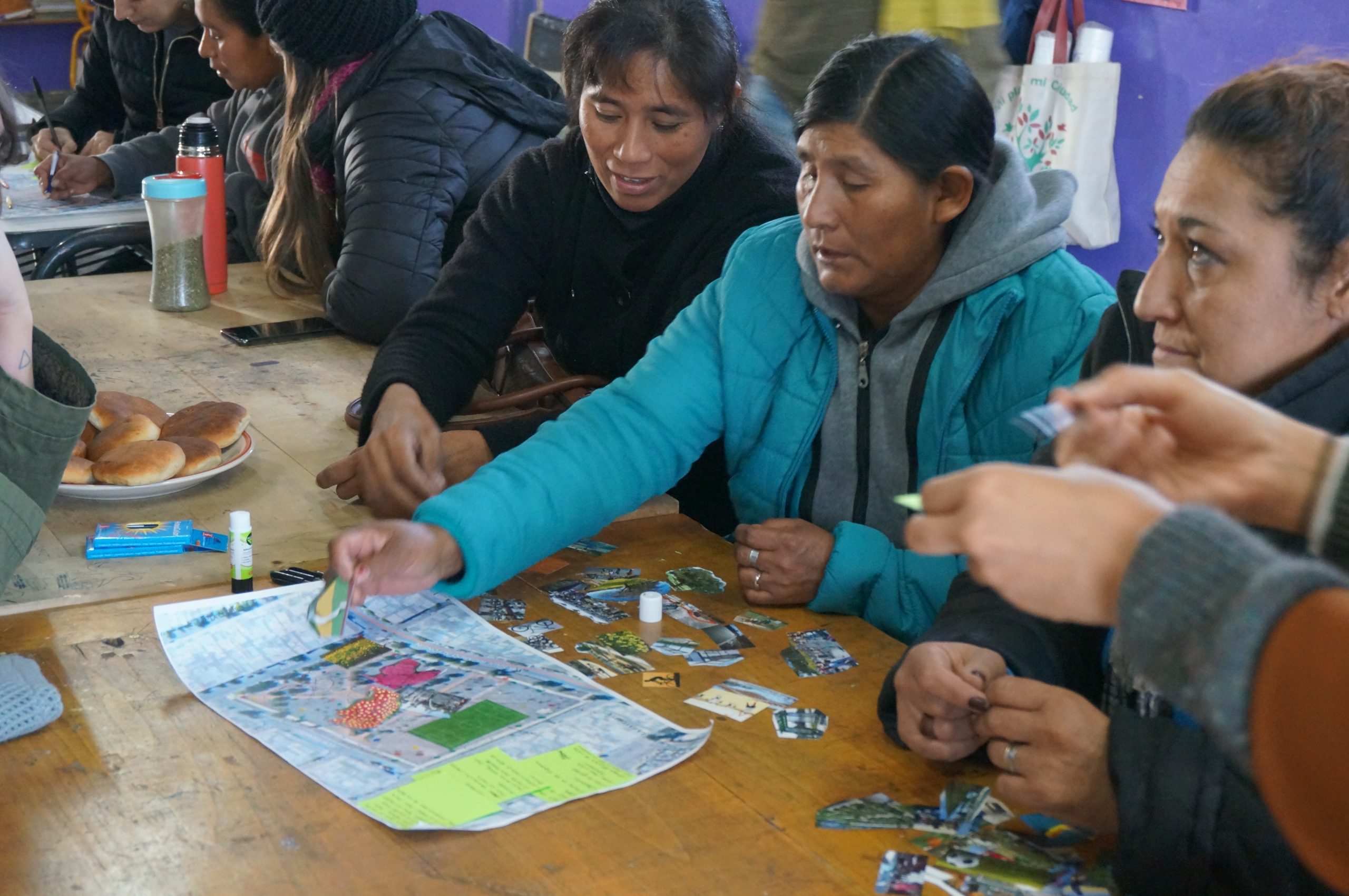A group of people, primarily women, are seated around a wooden table during a community workshop. They're actively collaborating on a large park design map, placing small pictures and icons onto the drawing. One woman in a bright blue jacket is reaching to place an item on the map.