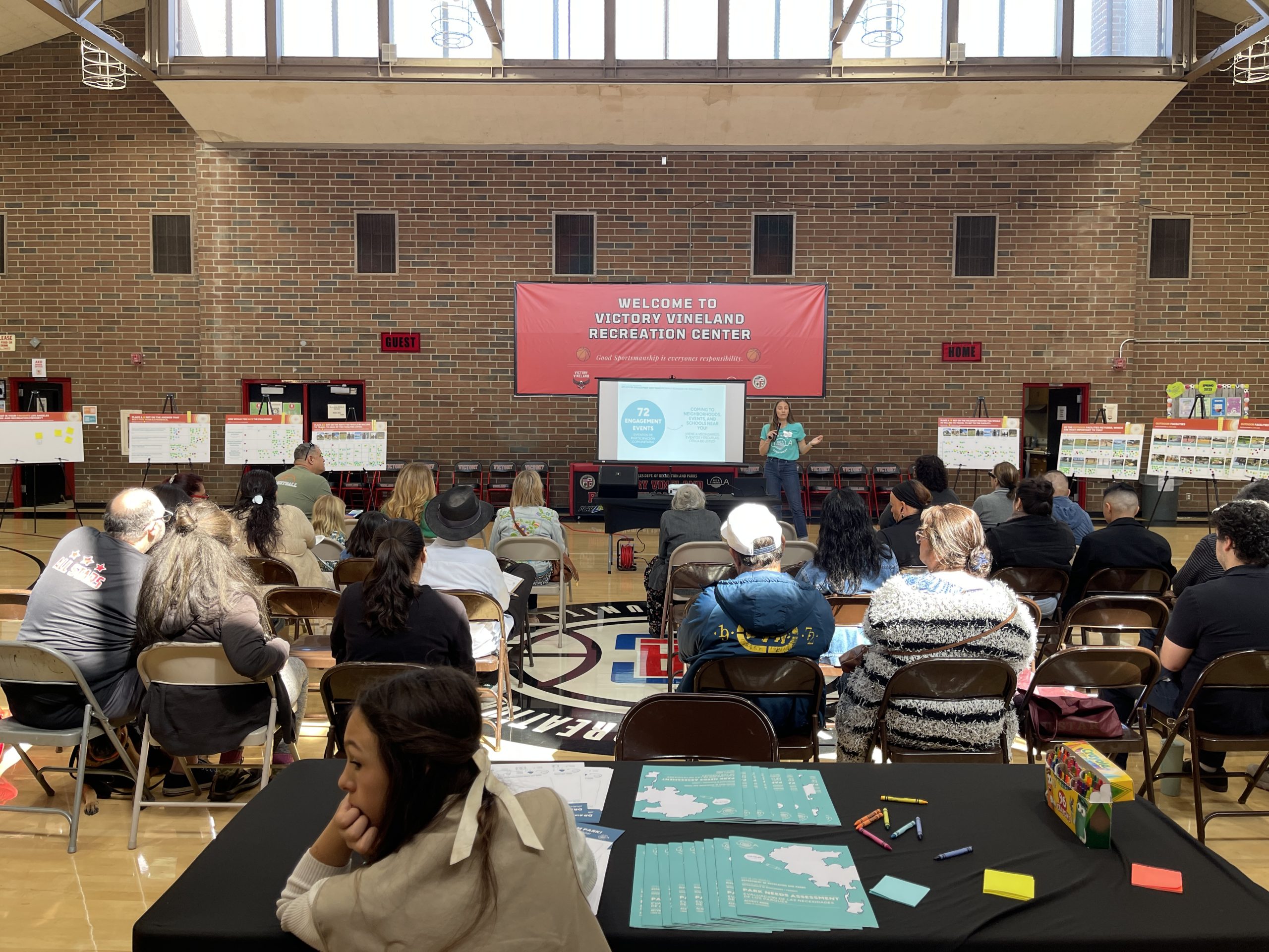 A community meeting is taking place indoors, in what appears to be a gymnasium. A woman stands in front of a projection screen and a large banner that reads, "WELCOME TO VICTORY VINELAND RECREATION CENTER." She is giving a presentation to a large audience seated in rows of folding chairs. Informational posters are also visible on easels on both sides of the room.