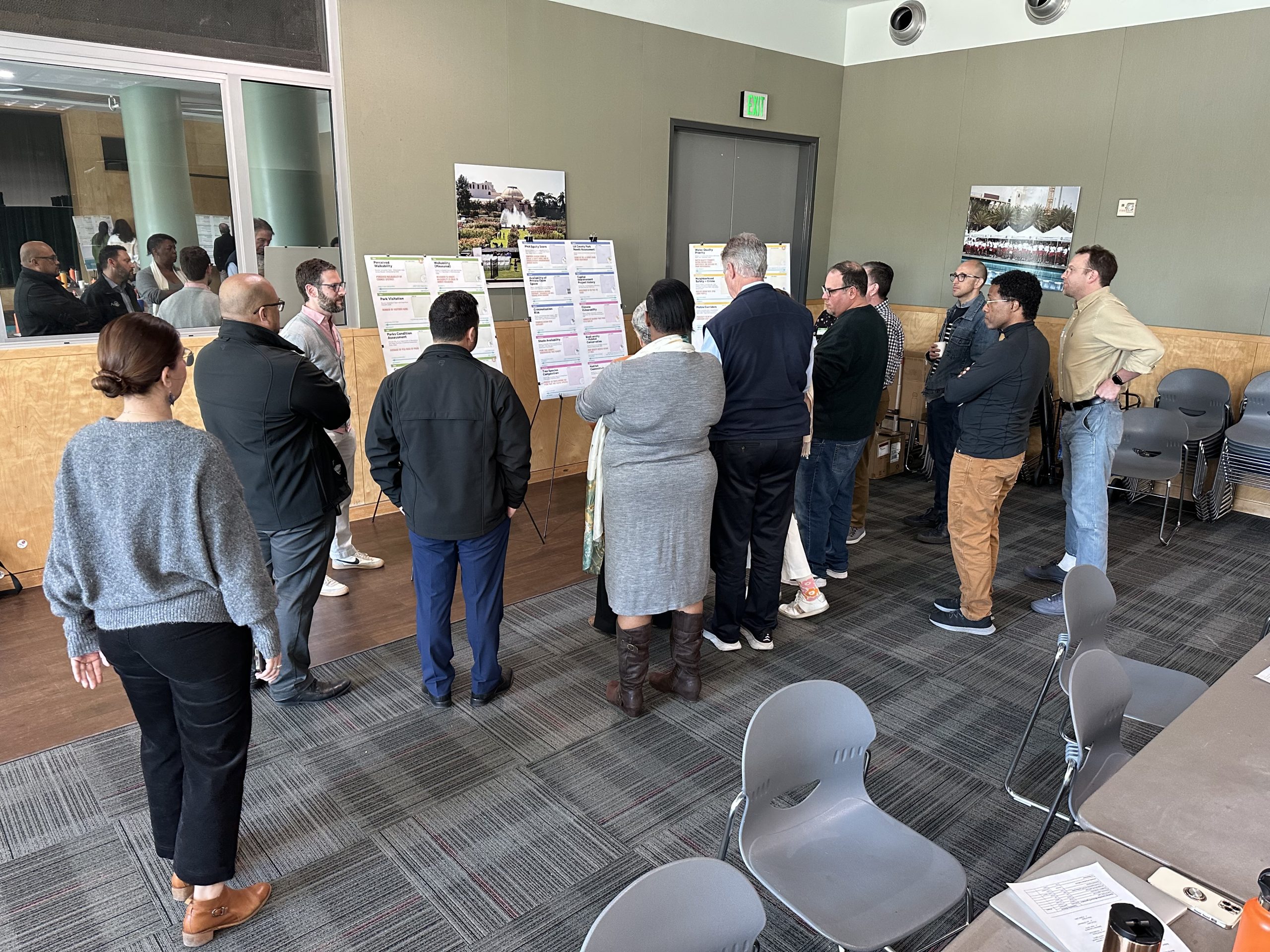 A group of people is gathered in a modern meeting room, standing and interacting with three large poster boards displayed on easels. The people are talking to one another while looking at the posters, which contain text and graphics, suggesting an engagement meeting.