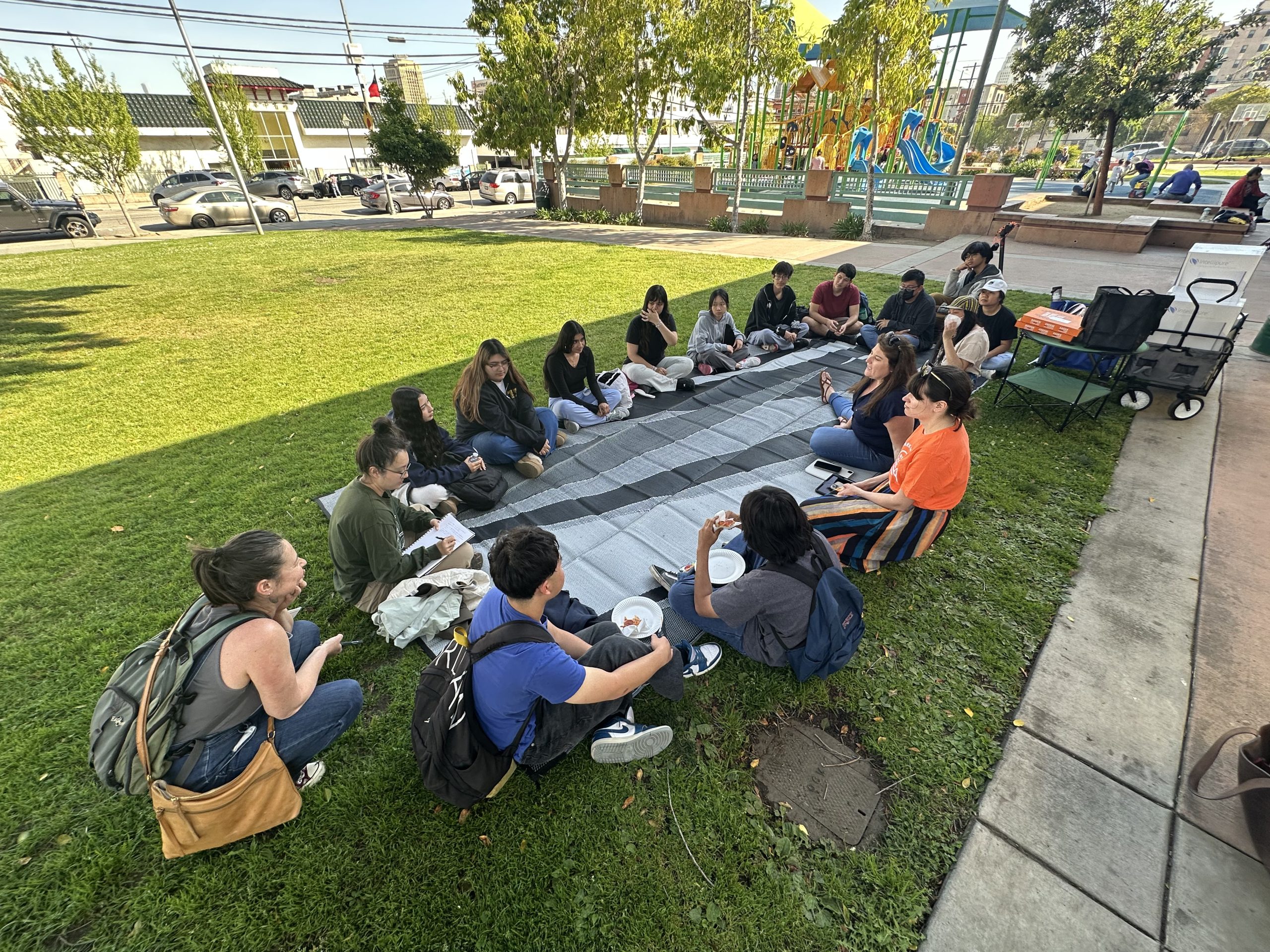 A woman leads an informal workshop for a group of young people seated in a circle on a striped blanket in a grassy park. A playground and a street are visible in the background.