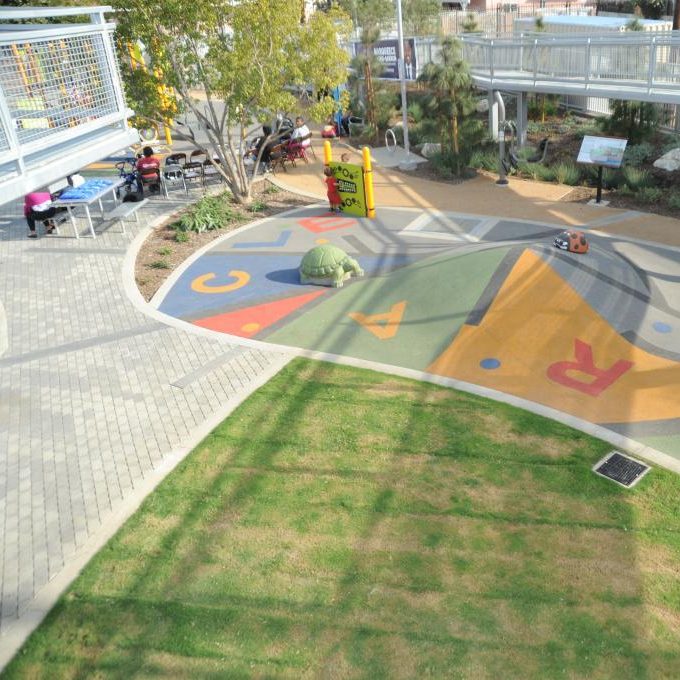 Outdoor park with a colorful floor mural, dirt pathways, greenery, educational signage, and elevated, white, metal walkways.