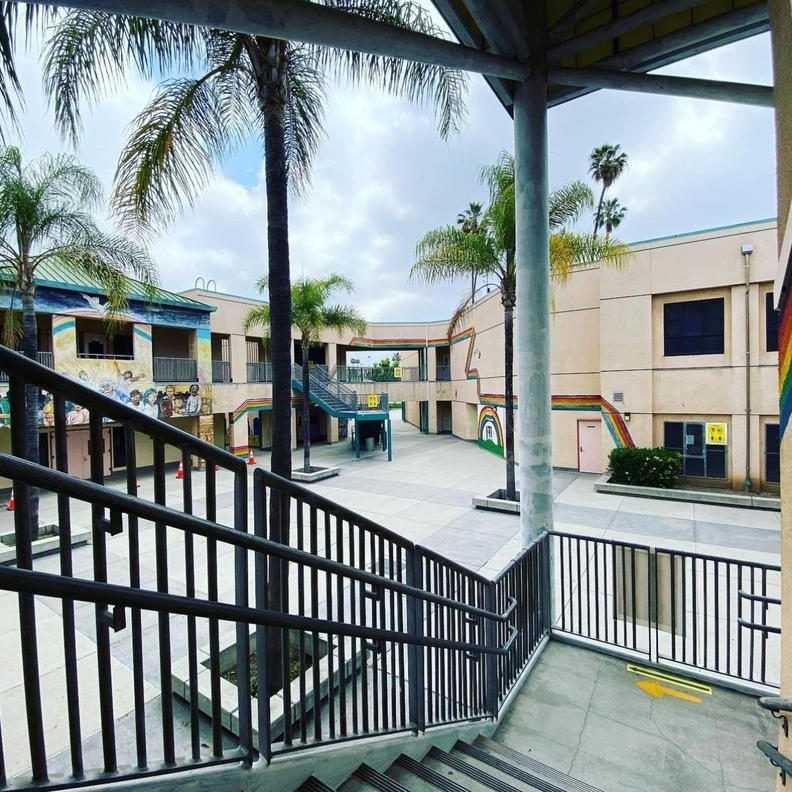 Outdoor courtyard of a two-story tan building with rainbows painted on the walls on a cloudy day. The area is empty save for four palm trees.
