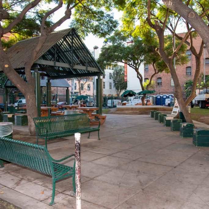 Outdoor seating area with green benches, picnic tables, and trees providing shade over the area.